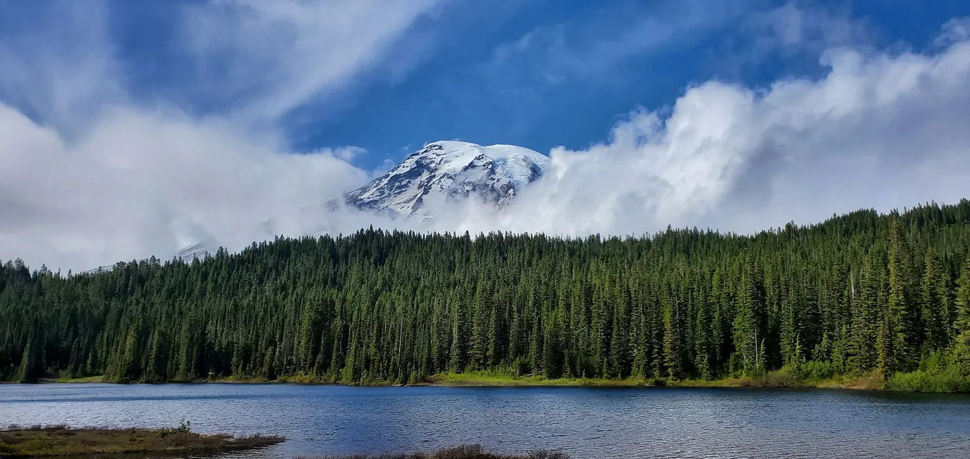 Mount Rainier shrugging off clouds above a treeline and lake